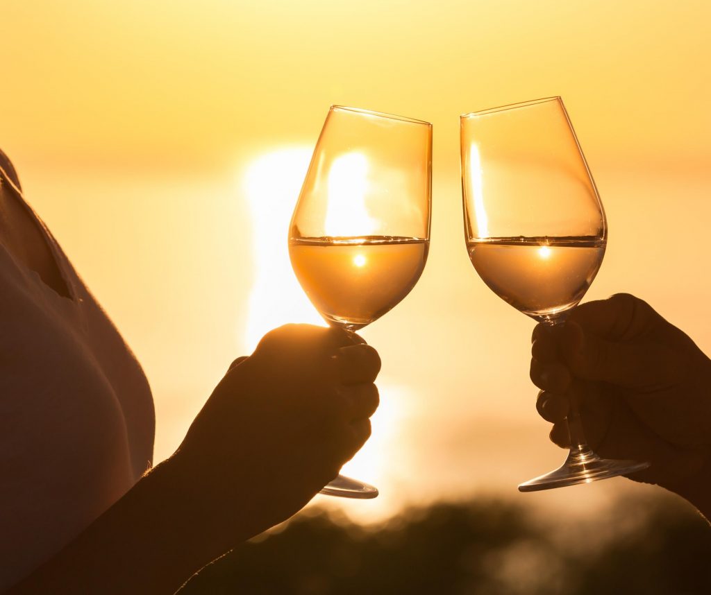 Close up of silhouette of two white wine glasses held and being clinked together with sun setting in background