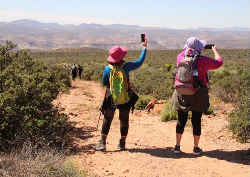 Two ladies on a hiking trail wearing sunhats with backpacks taking pictures with their smartphones of the Schaap River Canyon in the distance. In Namaqualand. The ladies are facing away from the camera. 