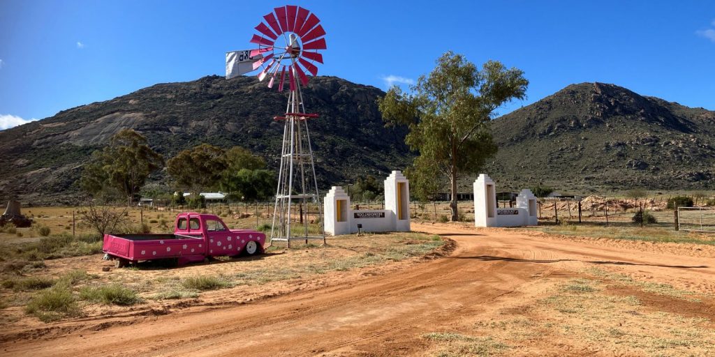 Photo of white farm gate with white brick pillar gates, a pink wind pump and tree in mid ground, hill in background and the chassy of an old pink car in foreground - in Namaqualand, South Africa
